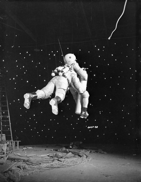 In this production still for "Destination Moon," two astronauts are suspended on wires. One holds a large gas canister. The other is grabbing the first. An unattached tethering line floats above them and to the right. Behind them are stars glowing in a black sky, below, bits and pieces of studio equipment including an unused safety net. Image ID: 120934 is an alternate setup for this shot that does not use wires.