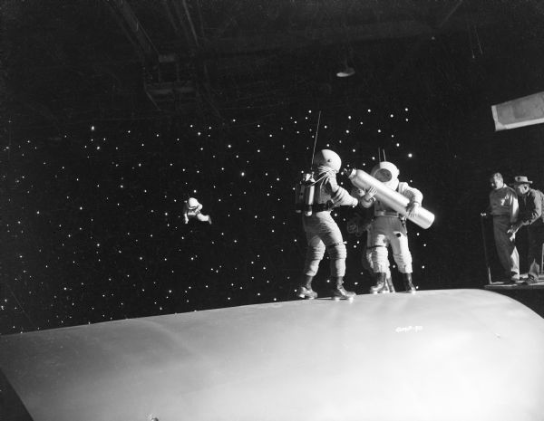 Three astronauts stand on the outer hull of a spaceship. One of them holds a large gas canister. Behind them, against a starry, black sky, floats a companion who has become untethered and is floating away from the ship. On the right, two technicians stand on a scaffold watching the scene.