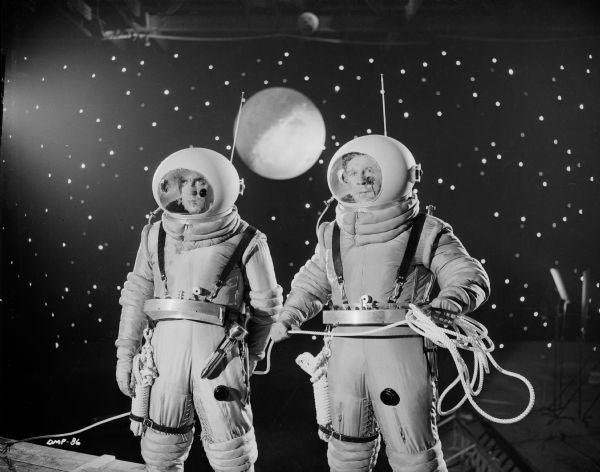 In a production still for "Destination Moon," two astronauts, played by Warner Anderson (left) and John Archer (holding rope) look off to the distance. Amid the stars in the background is the moon. In the lower left, lighting equipment can be seen.