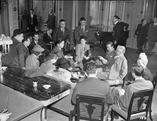Charlie Chaplin meets the press at the Waldorf-Astoria in New York on October 13, 1940, to promote "The Great Dictator." About fifteen reporters stand or sit on upholstered chairs listening to Chaplin. A baby grand piano is in the background.