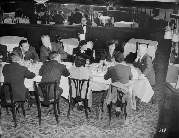 Charlie Chaplin talking to nine reporters sitting at the end of a long restaurant table at the Waldorf Astoria in New York. Behind them, a waiter is watching, and behind him are four large mirrors.