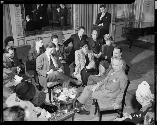 Charlie Chaplin talking to fifteen reporters sitting on upholstered chairs around him in a meeting room at the Waldorf Astoria in New York. A small coffee table is crowded with drinks.