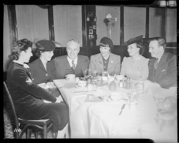 Charlie Chaplin is sitting drinking coffee with four women and a man in a restaurant in New York's Waldorf Astoria Hotel. The meeting is related to the New York premiere of "The Great Dictator." Everyone is smiling.