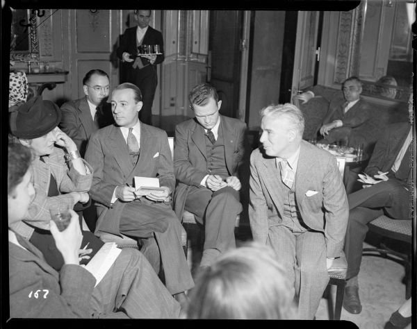 Charlie Chaplin listening to a woman reporter during a press conference at the Waldorf Astoria related to the New York premiere of "The Great Dictator." About eight reporters are sitting around Chaplin. In the background, a waiter is bringing more drinks.