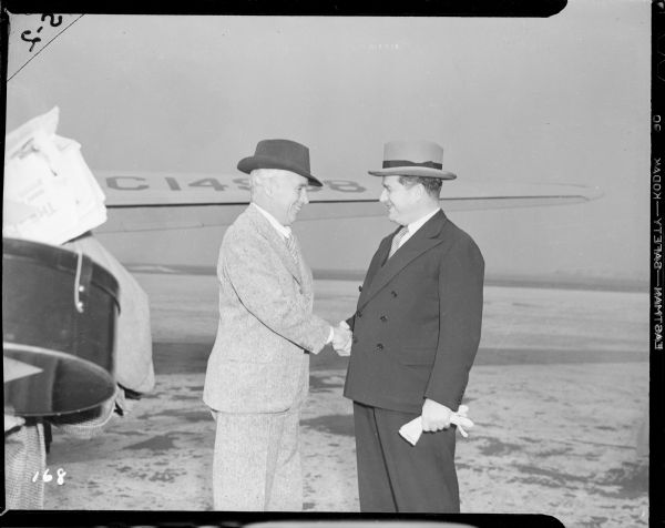 Charlie Chaplin grinning and shaking hands with a man in a dark suit. They are at an airfield next to a plane. Perhaps this is Chaplin's arrival for the New York premiere of "The Great Dictator."