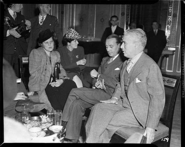 Charlie Chaplin sitting talking to reporters, about six of whom are visible. A small table covered with glasses of ice water, a coffee cup, cigarettes, etc., is at Chaplin's knee. Chaplin is at New York's Waldorf Astoria promoting "The Great Dictator."