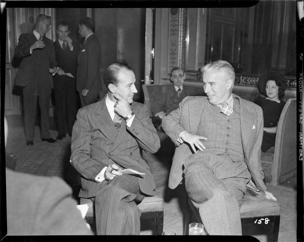 Charlie Chaplin and a reporter sitting side by side during a meeting of the press at the Waldorf Astoria for the New York premiere of "The Great Dictator." In the background two reporters are sitting and three are standing.