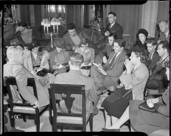 Charlie Chaplin is sitting on the far left with his arm draped over the chair and is talking to a circle of about twelve seated reporters during a meeting of the press at the Waldorf Astoria for the New York premiere of "The Great Dictator." In the background is a man with a press camera with a flash attachment.