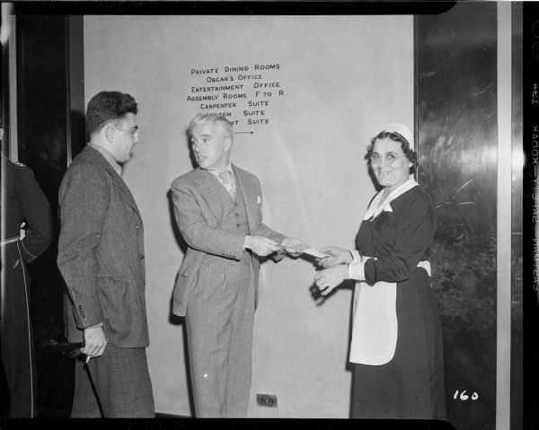 Charlie Chaplin talking over his shoulder to a man while handing a hotel maid a piece of paper which he has perhaps autographed. They are in a hallway of the Waldorf Astoria Hotel in New York.