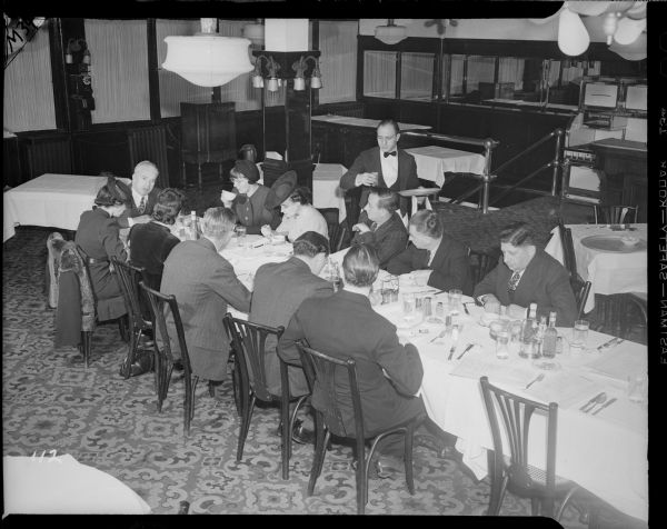 Charlie Chaplin sits at the far end of a long restaurant table at the Waldorf Astoria talking to ten reporters. A waiter serves drinks.