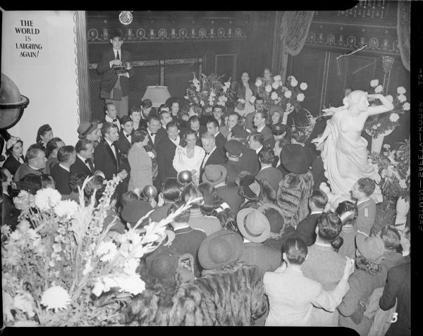 In the center of a large crowd of people in the Capitol Theatre, actress Paulette Goddard dressed in white smiles for a press photographer just a few feet in front of her. Left of her is Jack Oakie, on the right is Charlie Chaplin. Near the back wall, another press photographer stands on the furniture to get a better view. Near him "The World is laughing again!" is painted on the wall. It is the New York premiere of "The Great Dictator" on October 15, 1940.
