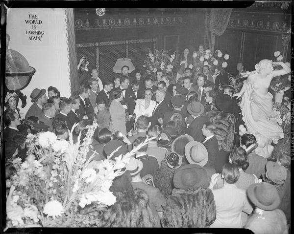 In the center of a large crowd of people in the lobby of the Capitol Theatre, actress Paulette Goddard dressed in white is smiling broadly as is Charlie Chaplin standing on the right. It is the New York premiere of "The Great Dictator" on October 15, 1940.