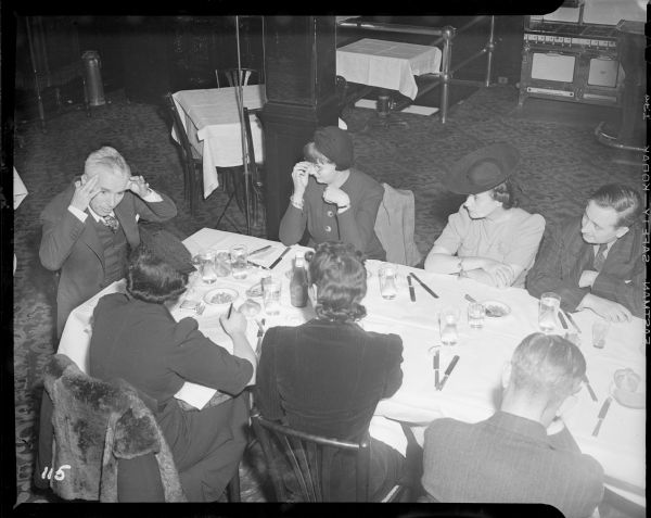 Charlie Chaplin is sitting at the far left end of a restaurant table gesturing with his hands held by his forehead. Six reporters are listening to him. This is a press conference at the Waldorf Astoria Hotel promoting the New York premiere of "The Great Dictator."