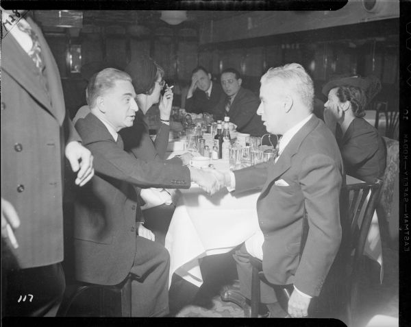Charlie Chaplin, sitting at the end of a long restaurant table, shaking hands with the man sitting on his left. Down the table are five or six more reporters. This is a press conference held in the Waldorf Astoria for the New York premiere of "The Great Dictator."
