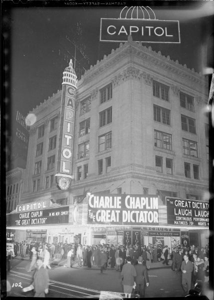 Crowds gathering under the marquee of the Capitol Theatre for the New York premiere of Charlie Chaplin's "The Great Dictator" the evening of October 15, 1940. The photograph was taken catercorner from the theater at the intersection of West 51st Street and Broadway with pedestrians in the foreground. In addition to the marquee, the photograph shows the long vertical sign hanging from the side of the building and the sign on the roof six stories up.