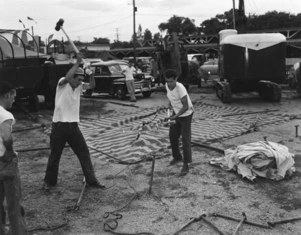 Two men are pounding a tent stake into the ground at the State Fair Park. Behind them are excavating machinery, automobiles and permanent structures associated with the Wisconsin Centennial Exposition.