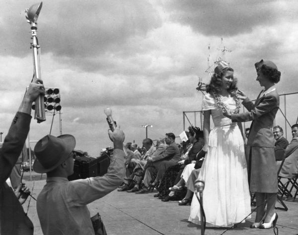 A woman in a uniform places a lei of orchids around Margaret McGuire's neck as she accepts the role of Centennial Queen on a stage at the Wisconsin Centennial Exposition, she was also the hostess at the Dairy Pavillion. Margaret McGuire is the 1948 (first) Alice in Dairyland. Men at the foot of the stage can be seen taking photographs with flash-bulbs attached to their cameras.