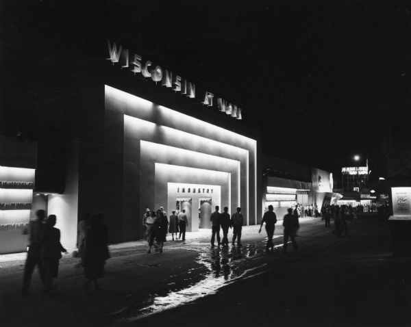 Night view of small crowd of people gathered at outside entrance of the "Wisconsin At Work" building at the Wisconsin Centennial Exposition.
