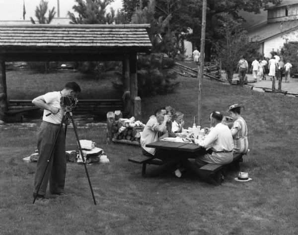 A man films a family as they sit at a picnic table and eat lunch at the Wisconsin Centennial Exposition.