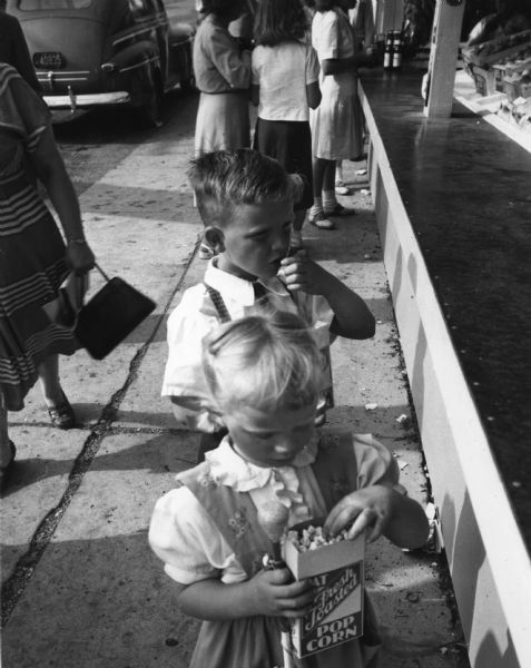A young boy and girl are eating popcorn from a box. They are standing next to the snack counter at the Wisconsin Centennial Exposition.