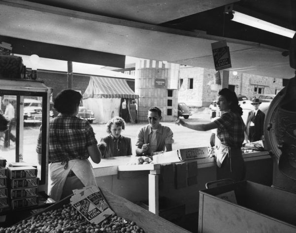 View from interior of a man wearing sunglasses, and a woman at the snack counter at the Wisconsin Centennial Exposition. Two women are behind the counter serving them. Empty and flattened boxes for popcorn are seen on top of a large box of peanuts.