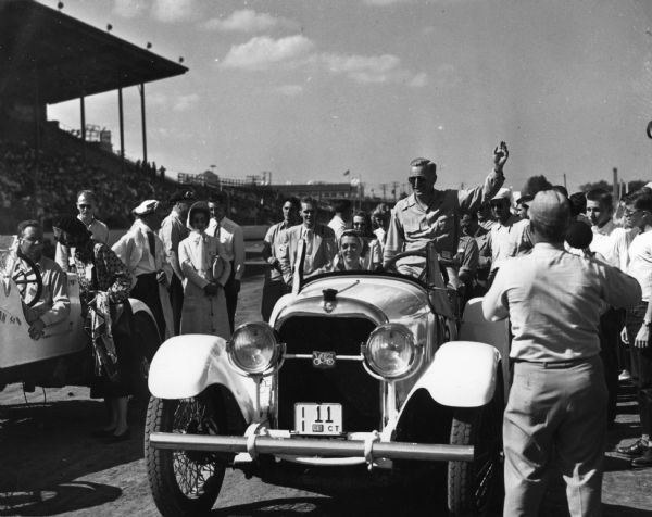 A man surrounded by a small crowd waves from the seat of an automobile in front of the Wisconsin Centennial Exposition grandstand.