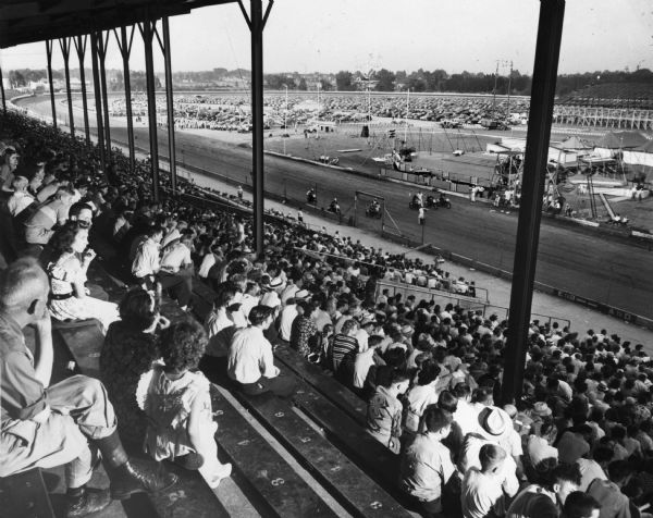 View from behind of crowd at the Wisconsin Centennial Exposition grandstand watching motorcycles line up before a race. In the background are tents as well as a number of parked cars.