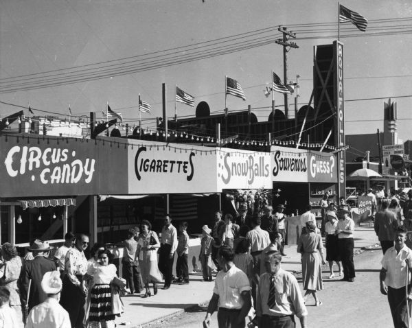 Crowded midway at the Wisconsin Centennial Exposition. Signs above booths read "Circus Candy," "Cigarettes," "Snow Balls," "Souvenirs," "Guess?" and "Aqua Follies."