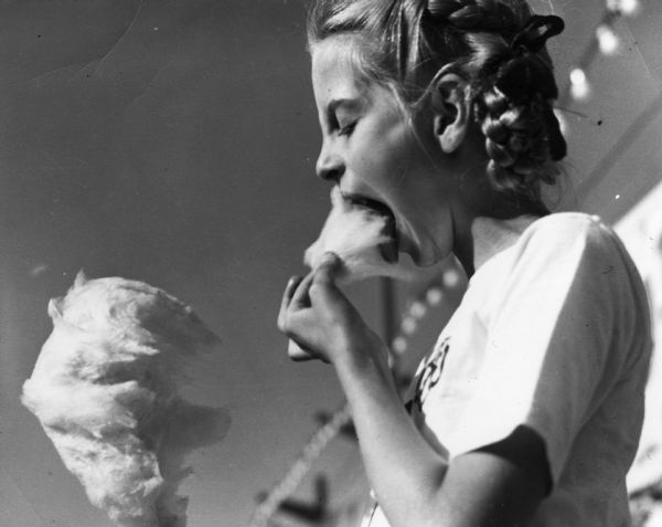 Close-up of a young girl at the Wisconsin Centennial Exposition eating cotton candy. There is a string of lights in the background.