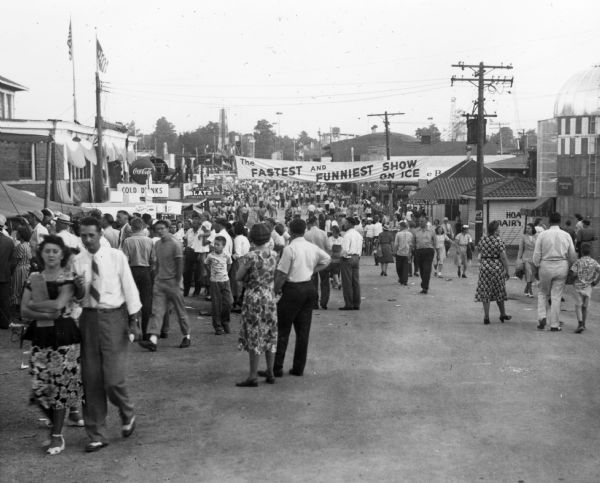 A crowded street at the Wisconsin Centennial Exposition State Fairgrounds. Above the crowd is a banner reading "The Fastest And Funniest Show On Ice." American flags and utility lines are visible as well as a small silo on the right.