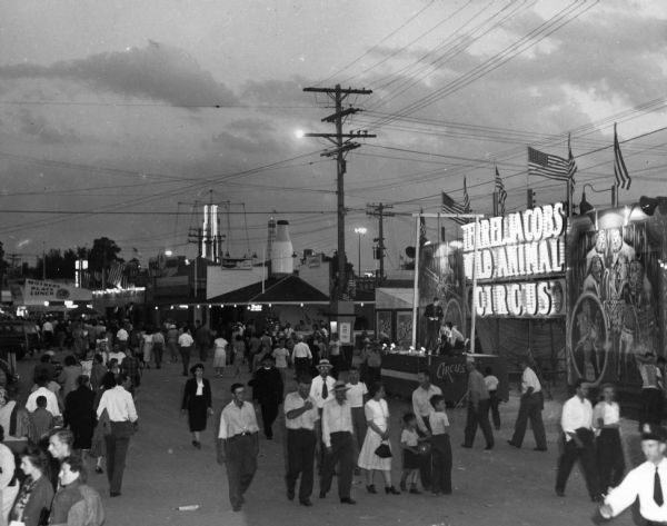 A crowd walking the grounds of the Wisconsin Centennial Exposition in the early evening. An electric sign on the right says "Terrel Jacobs Wild Animal Circus," and in the background is a sign for "Mother's Plate Lunch."