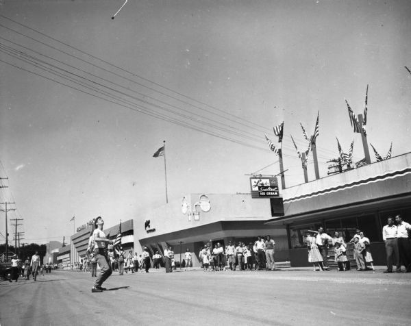 A crowd watches a young man throwing a baton into the air at the Wisconsin Centennial Exposition. Behind him is the "Alice In Dairyland" building, featuring a neon sign of a pitcher of milk filling a glass, as well as other permanent fair-related buildings.
