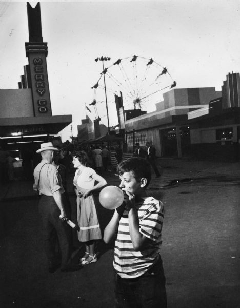 A young boy inflates a balloon at the Wisconsin Centennial Exposition. Three people stand behind him near the "Merry Go Round" building, and in the background a crowd is walking near booths and a Ferris Wheel on the midway.