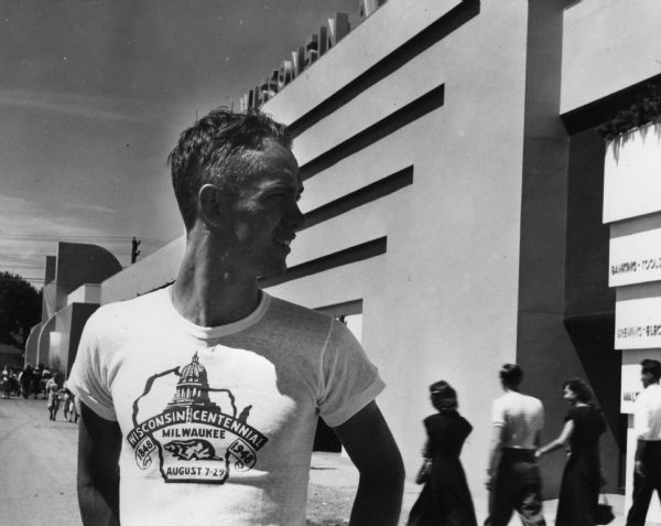 A man wearing a "Wisconsin Centennial, Milwaukee, August 7-29" t-shirt stands in front of the "Wisconsin At Work" building at the Wisconsin Centennial Exposition. The shirt features the Wisconsin state capitol dome and a badger in an outline of the state.