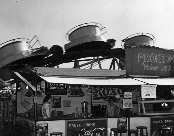 Exterior view of a "Pronto-Dog" stand at the Wisconsin Centennial Exposition staffed by three people. Behind the stand is an amusement park ride.