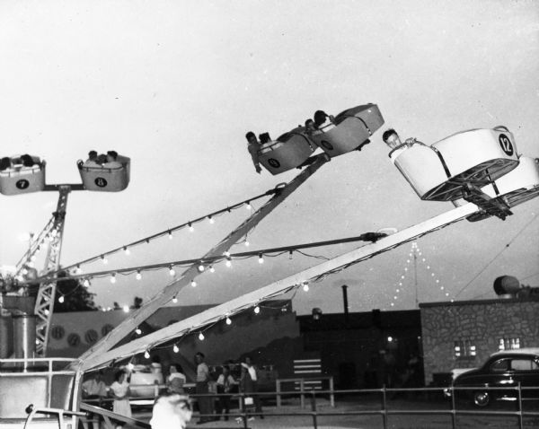 A teenage boy caught looking directly at the photographer while riding an amusement park ride at the Wisconsin Centennial Exposition midway in the early evening twilight. In the background is the "Bubble Bounce."