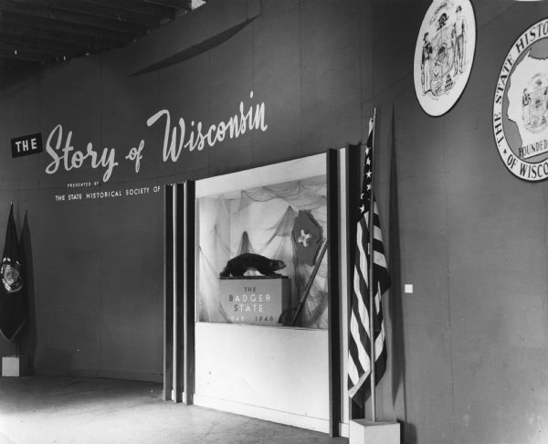 Display at entrance of The Wisconsin State Historical Society's "The Story of Wisconsin" exhibit at the Wisconsin Centennial Exposition. The display prominently features a stuffed badger, as well as the flag of the United States, the state flag and state seals.