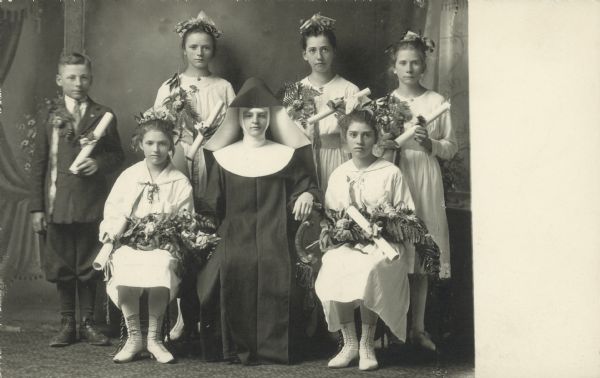Photographic postcard of a full-length studio portrait of a nun, one young man and five young women in front of a painted backdrop. The children are all holding scrolls, possibly a diploma, and the girls have bouquets. The girls are all dressed in white. 

