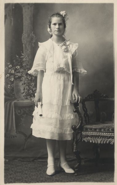 Photographic postcard of a full-length studio portrait of a Young woman in front of a painted backdrop. She is standing posed next to a chair with a scroll in her hand, possibly a diploma. The girl is wearing all white and has flowers in her hair. She is also wearing a corsage.