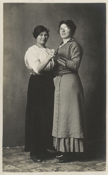 Photographic postcard of a full-length studio portrait of two women wearing dresses in front of a painted backdrop. They are facing each other and holding hands as if they were about to dance.