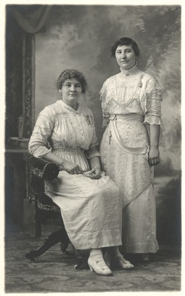 Photographic postcard of a full-length studio portrait of two women in front of a painted backdrop. One woman is seated and is one standing, and they are wearing fashionable dresses. Both women are wearing lapel watches pinned to their bodices.