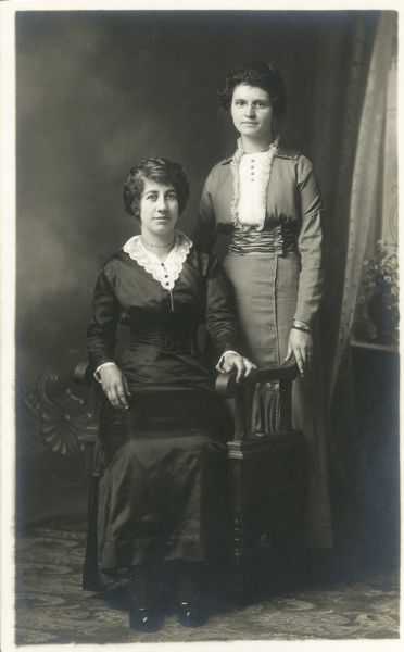 Full-length studio portrait of two women, one seated and one standing in front of a painted backdrop. They are both wearing fashionable dresses. Their names are Mrs. Joe Ziegler, on the left, and Mrs. Dridrich, on the right.