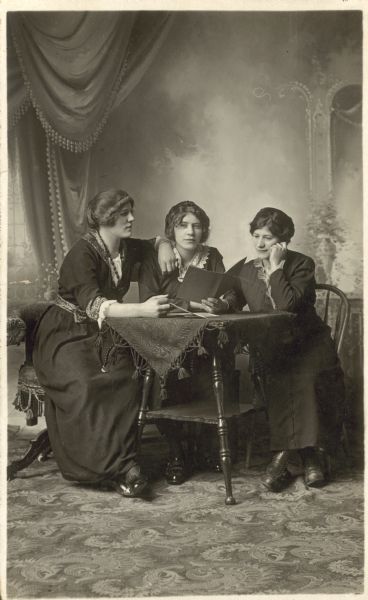 Full-length studio portrait of three women gathered around a table in front of a painted backdrop. One woman rests her forearm on her friends shoulder and another is supporting her cheek on her hand. They appear to be looking at a book.