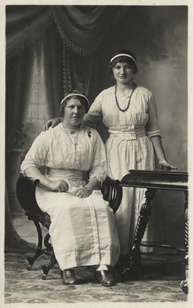 Full-length studio portrait of two women in front of a painted backdrop. One woman is seated and one is standing. One of the women is Annie Karls Dohm.