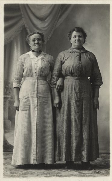 Full-length studio portrait in front of a painted backdrop of two women, with arms linked, standing in front of a painted backdrop.