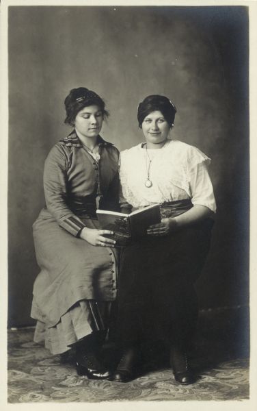 Full-length studio portrait of two women sitting in front of a painted backdrop sharing a book. Both women are wearing hair combs. One woman is looking at the camera.