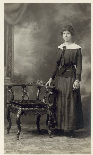 Full-length studio portrait in front of a painted backdrop of a woman dressed in a dark dress with a white collar and dark scarf. She stands next to an ornate chair.