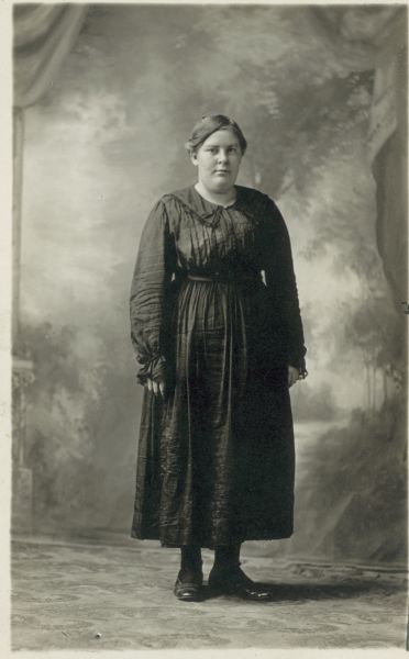 Full-length studio portrait of a standing woman dressed in a dark dress in front of a painted backdrop.