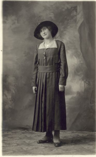 Full-length studio portrait of Isabel Berg Ripon, standing in a dark dress with with a light collar and a dark hat in front of a painted backdrop. She is wearing spats over her shoes.