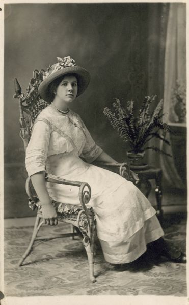 Full-length studio portrait of a woman seated in an ornate wicker chair in front of a painted backdrop. She is wearing a light-colored dress, pearls and a straw hat trimmed with flowers. A Boston fern is sitting on a table on her left.